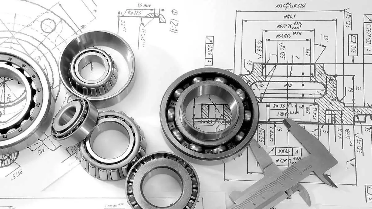Precision mechanical parts and ball bearings placed on technical engineering drawings, alongside a vernier caliper for dimension checking.
